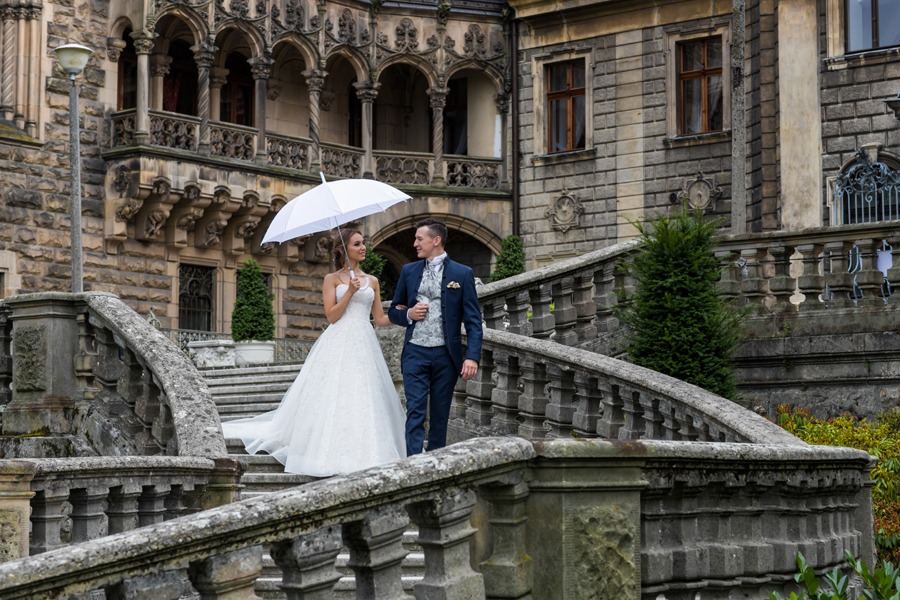 Para młoda, trzymająca biały parasol, schodzi po kamiennych schodach zamku w Mosznej koło Rybnika, w tle widoczne zdobienia architektoniczne.