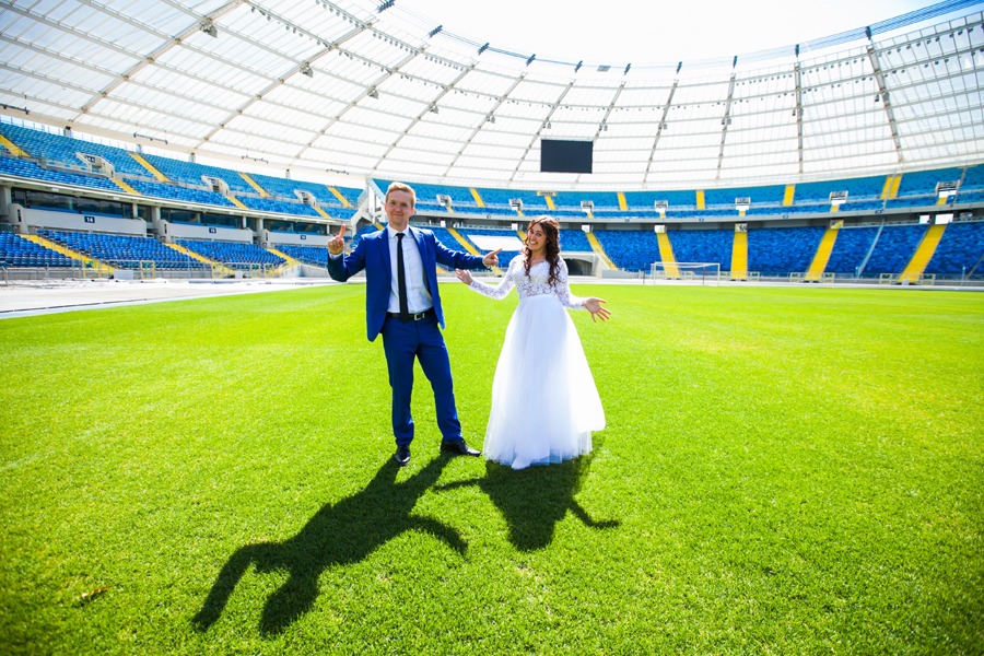 Para młoda w strojach ślubnych pozuje na zielonej murawie stadionu Śląskiego w Chorzowie, w tle widoczne niebieskie trybuny pod białym zadaszeniem.