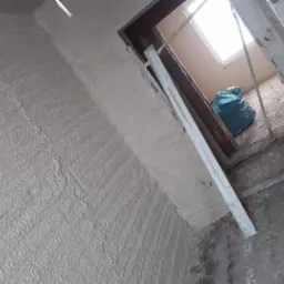 Świeżo nałożone tynki maszynowe z wyraźną fakturą na ścianie w trakcie prac wykończeniowych, widoczne elementy ościeżnicy i worki z odpadami.