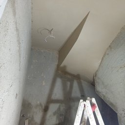 Tynki maszynowe - Wnętrze w trakcie prac wykończeniowych: surowe ściany, świeżo położone gładzie na suficie z wystającymi przewodami elektrycznymi, drabina malarska w rogu pomieszczenia.