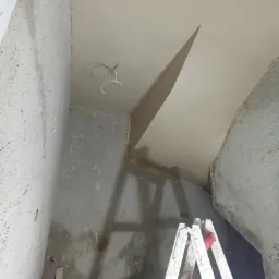 Wnętrze w trakcie prac wykończeniowych: surowe ściany, świeżo położone gładzie na suficie z wystającymi przewodami elektrycznymi, drabina malarska w rogu pomieszczenia.