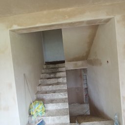 Tynki maszynowe - Surowe schody betonowe w trakcie prac wykończeniowych, ściany z widocznymi śladami tynku, niebieska miotła i wiadro z plandeką przy podstawie schodów.