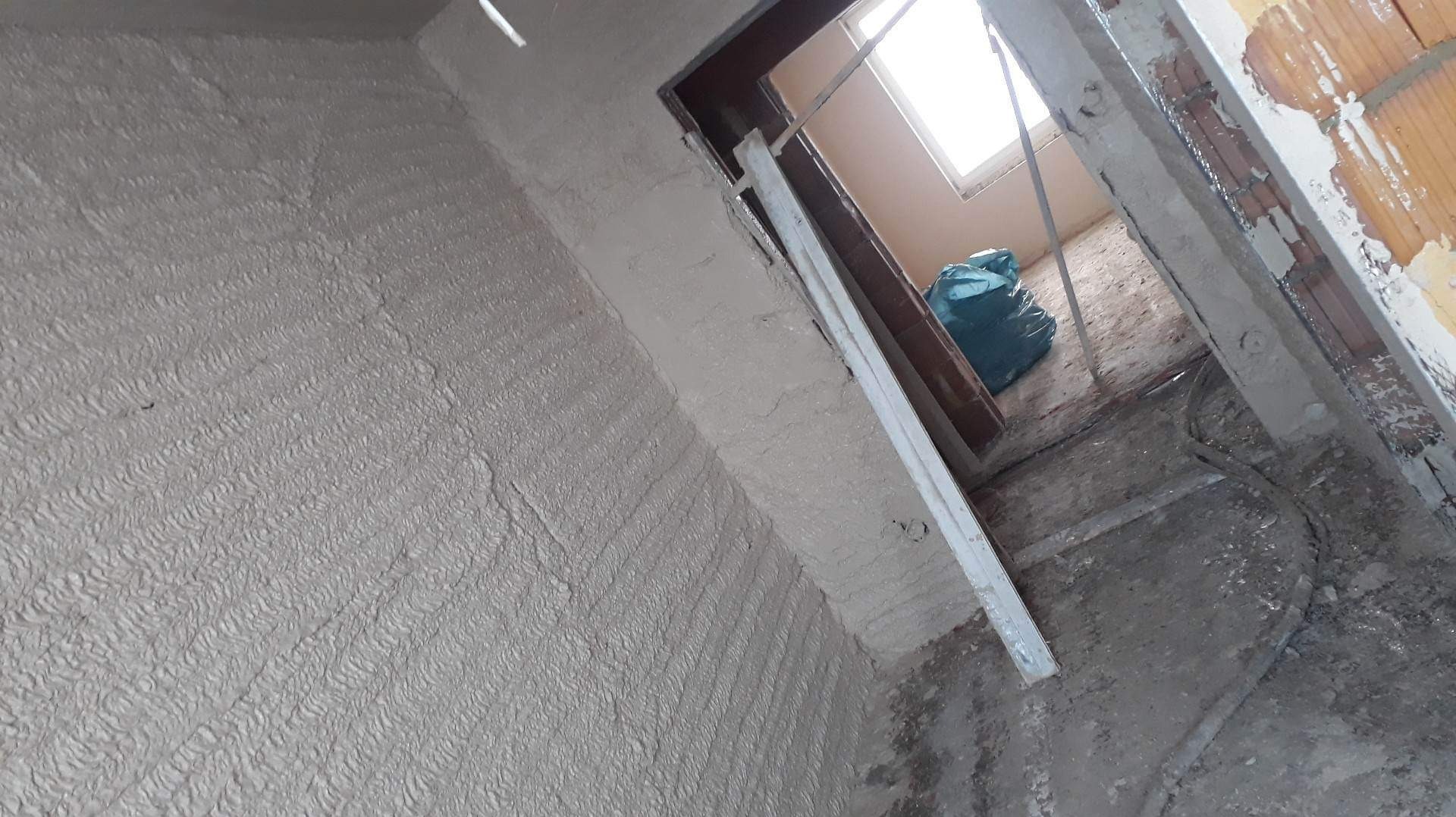 Świeżo nałożone tynki maszynowe z wyraźną fakturą na ścianie w trakcie prac wykończeniowych, widoczne elementy ościeżnicy i worki z odpadami.