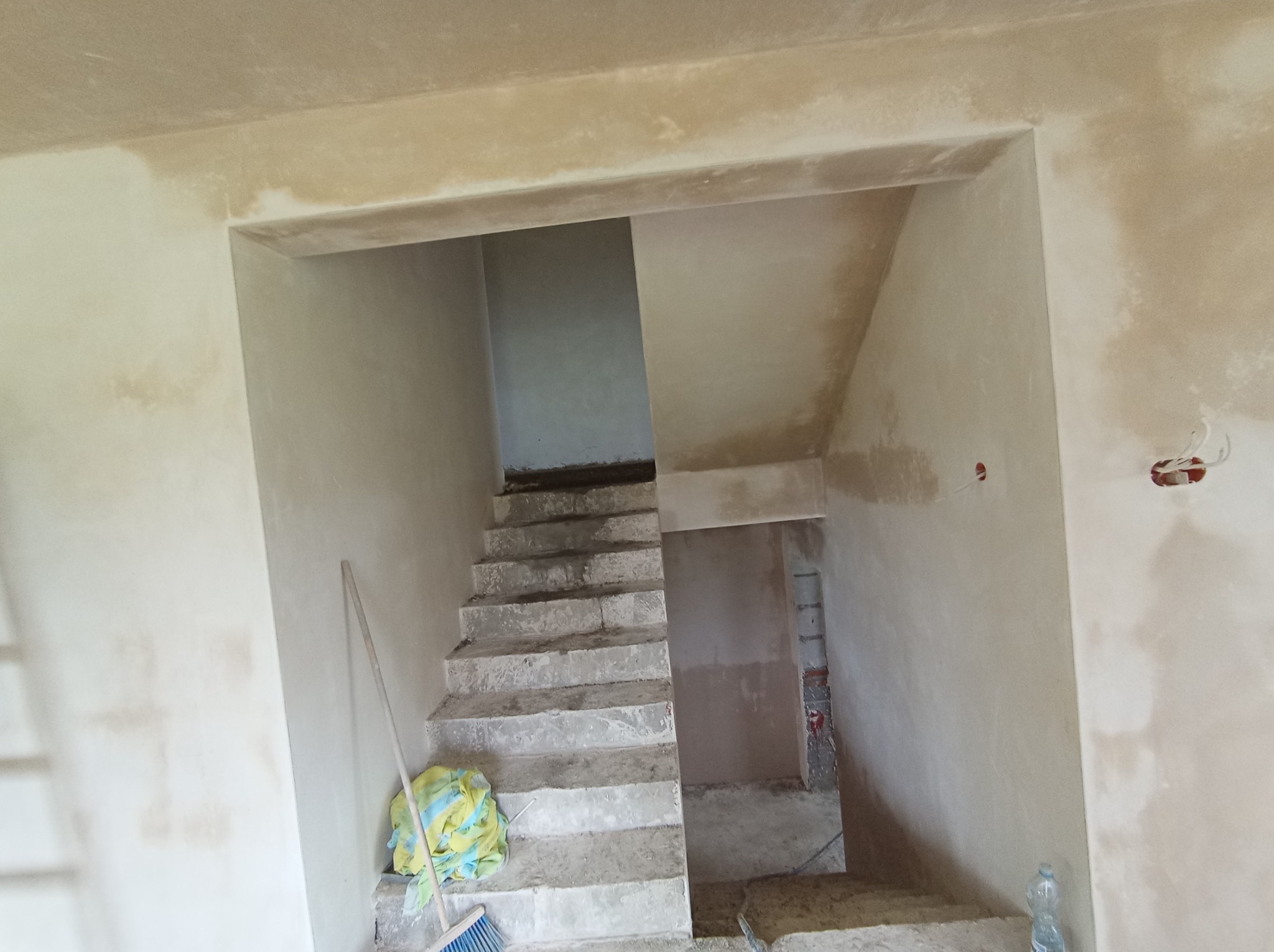 Surowe schody betonowe w trakcie prac wykończeniowych, ściany z widocznymi śladami tynku, niebieska miotła i wiadro z plandeką przy podstawie schodów.