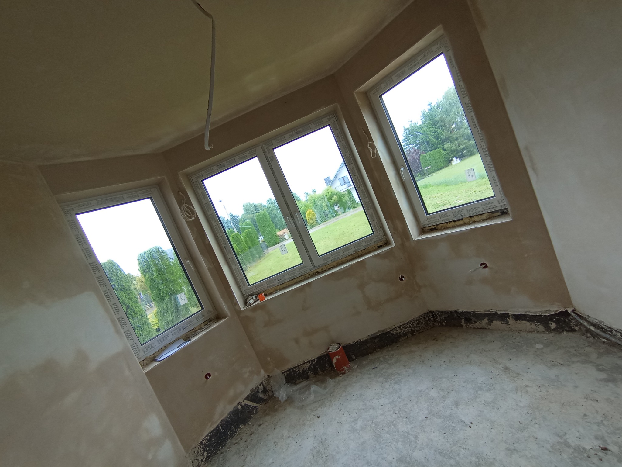 Wnętrze pokoju po tynkowaniu maszynowym, widoczne trzy okna z widokiem na zieleń, ściany w surowym tynku, podłoga przygotowana do dalszych prac wykończeniowych, wystające kable elektryczne z sufitu.