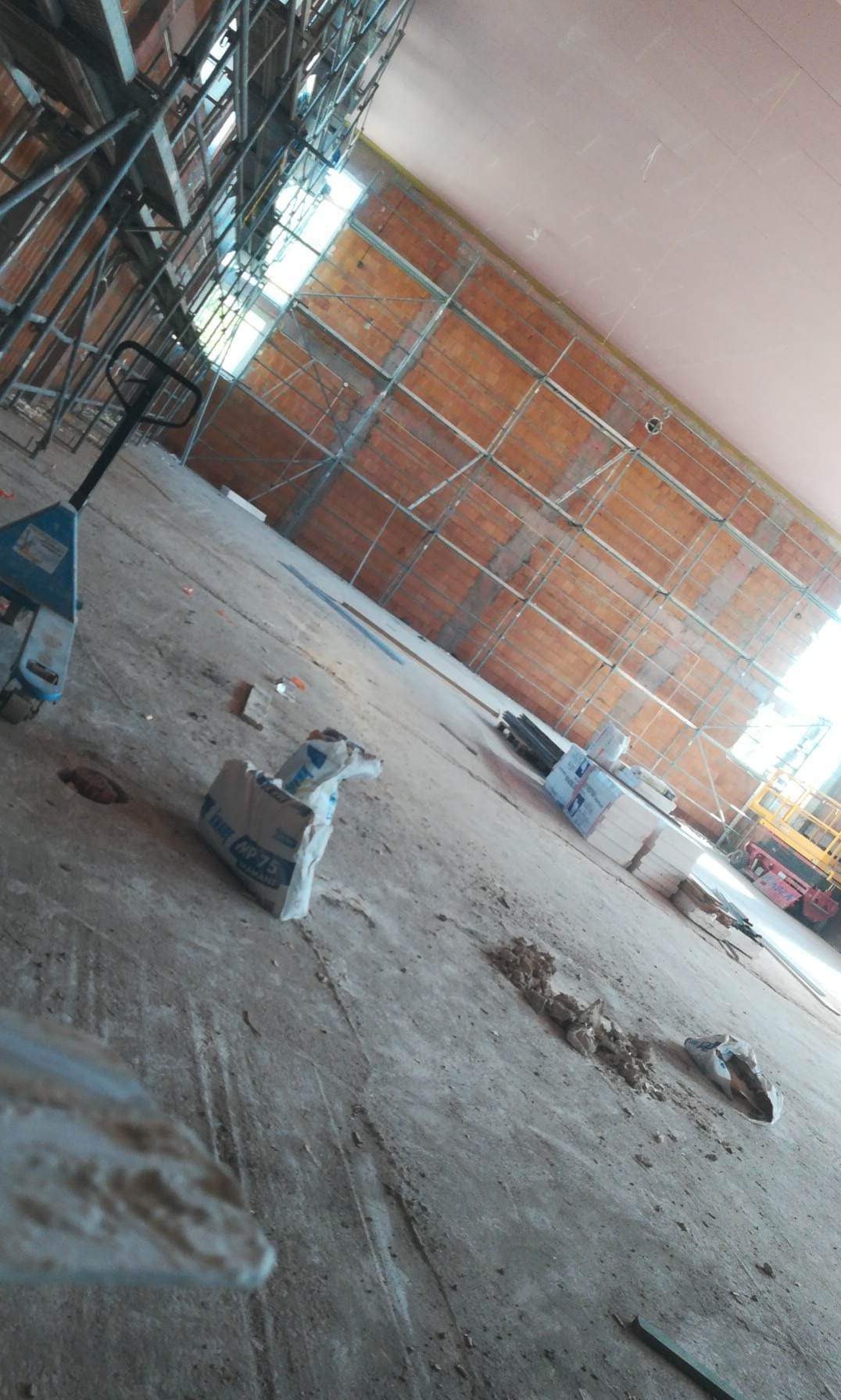 Wnętrze w trakcie prac wykończeniowych: widoczne rusztowania, ceglana ściana, worki z zaprawą i paleciak na betonowej podłodze.