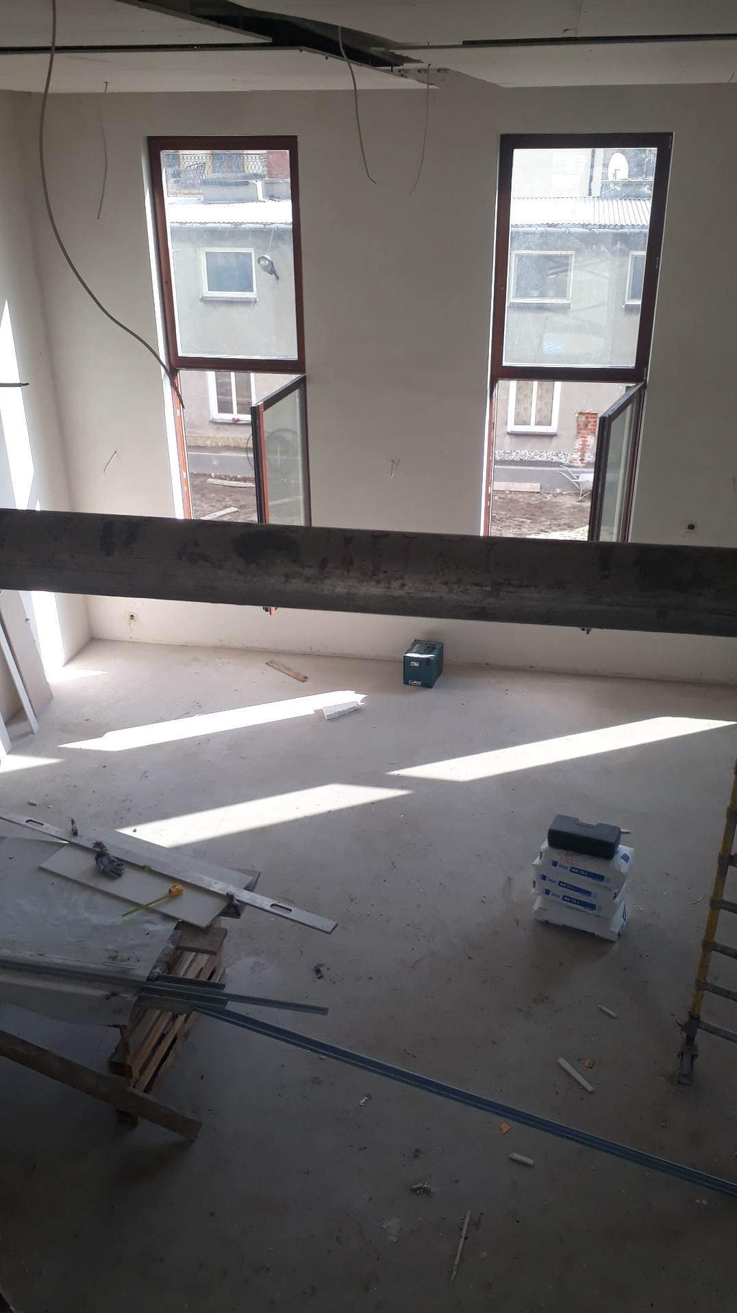 Surowe wnętrze w trakcie remontu z dwoma otwartymi oknami, widoczne materiały budowlane i narzędzia, surowy beton na podłodze, przygotowanie do prac wykończeniowych.