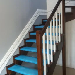 Drewniane schody z białymi balustradami, zabezpieczone niebieską folią ochronną podczas montażu, widok z dołu na jasnoszarą ścianę i fragment ściany wykończonej imitacją cegły.