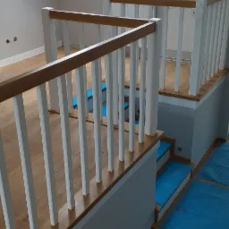 Jasne wnętrze z drewnianą antresolą i schodami, zabezpieczonymi niebieską folią ochronną, widok z góry na balustradę z białymi tralkami i drewnianym poręczą, w tle okna dachowe.