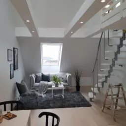 Jasne poddasze z oknem dachowym, szarą sofą, spiralnymi schodami i drewnianą drabiną malarską, po remoncie.