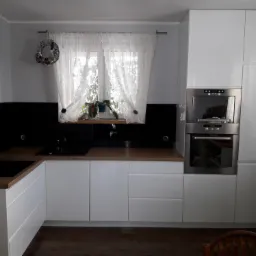 Nowoczesna kuchnia z białymi, lakierowanymi frontami szafek, drewnianym blatem i czarnym panelem nad blatem. Na oknie wisi wieniec i firanka.