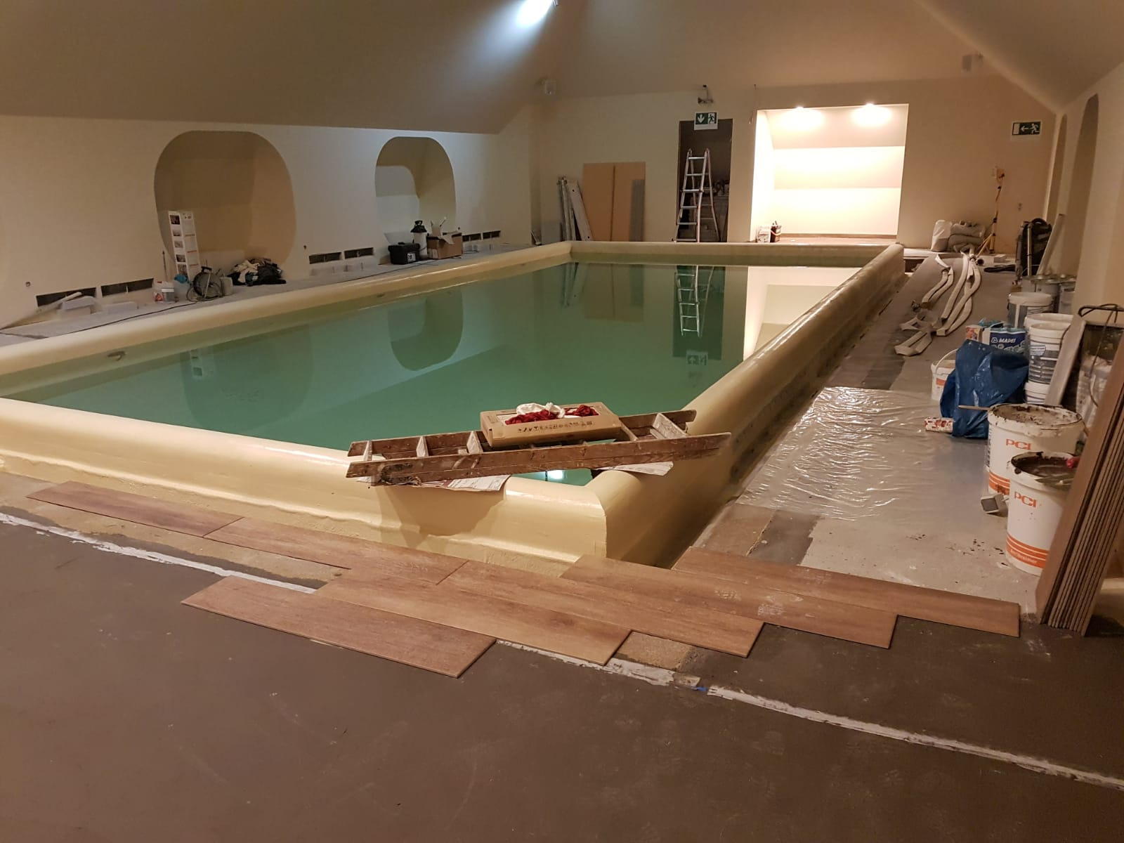 Wnętrze basenu w trakcie prac wykończeniowych, z widocznymi płytkami imitującymi drewno układanymi na posadzce wokół niecki, oraz narzędziami i materiałami budowlanymi w tle.