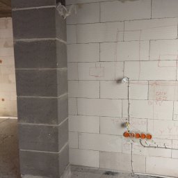 Twój Instalator Piotr Paluch - Ściana z betonu komórkowego z instalacją elektryczną w trakcie montażu: puszki pod gniazdka i przewody. Na ścianie widoczne szkice rozplanowania instalacji.
