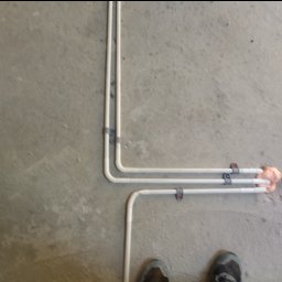 Twój Instalator Piotr Paluch - Białe rury przymocowane do szarego, surowego betonu klamrami. Rury zabezpieczone pianką montażową. Widoczne buty osoby wykonującej prace.