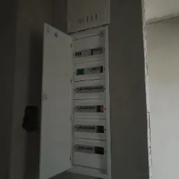 Otwarta skrzynka elektryczna z bezpiecznikami w surowym wnętrzu, widoczne przewody. Instalacja w trakcie prac wykończeniowych, ściany w stanie deweloperskim.