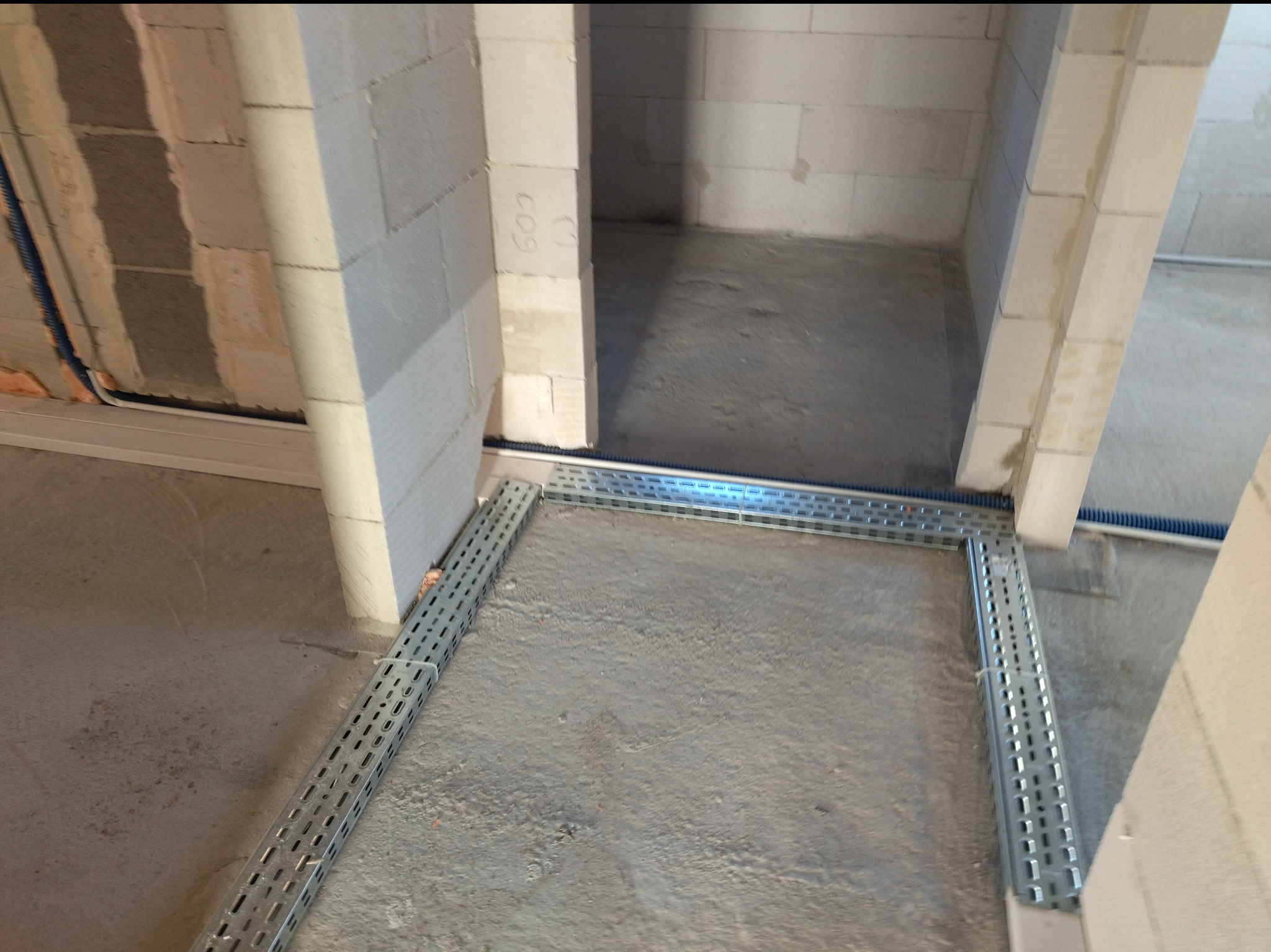 Wnętrze w trakcie budowy: ściany z betonu komórkowego, koryto kablowe przy podłodze, niebieska rura instalacyjna wzdłuż ściany. Widok z góry na surową przestrzeń.