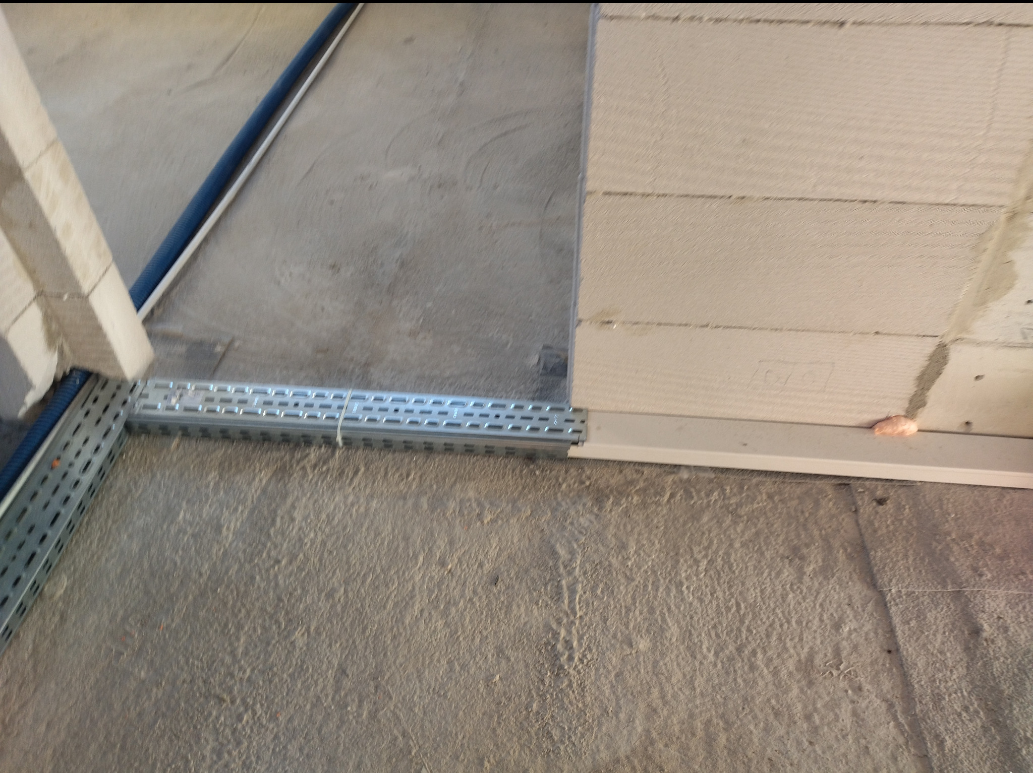 Szara, perforowana korytka kablowa zamontowana przy ścianie z betonu komórkowego. W pobliżu widoczna niebieska rura. Etap prac instalacyjnych.