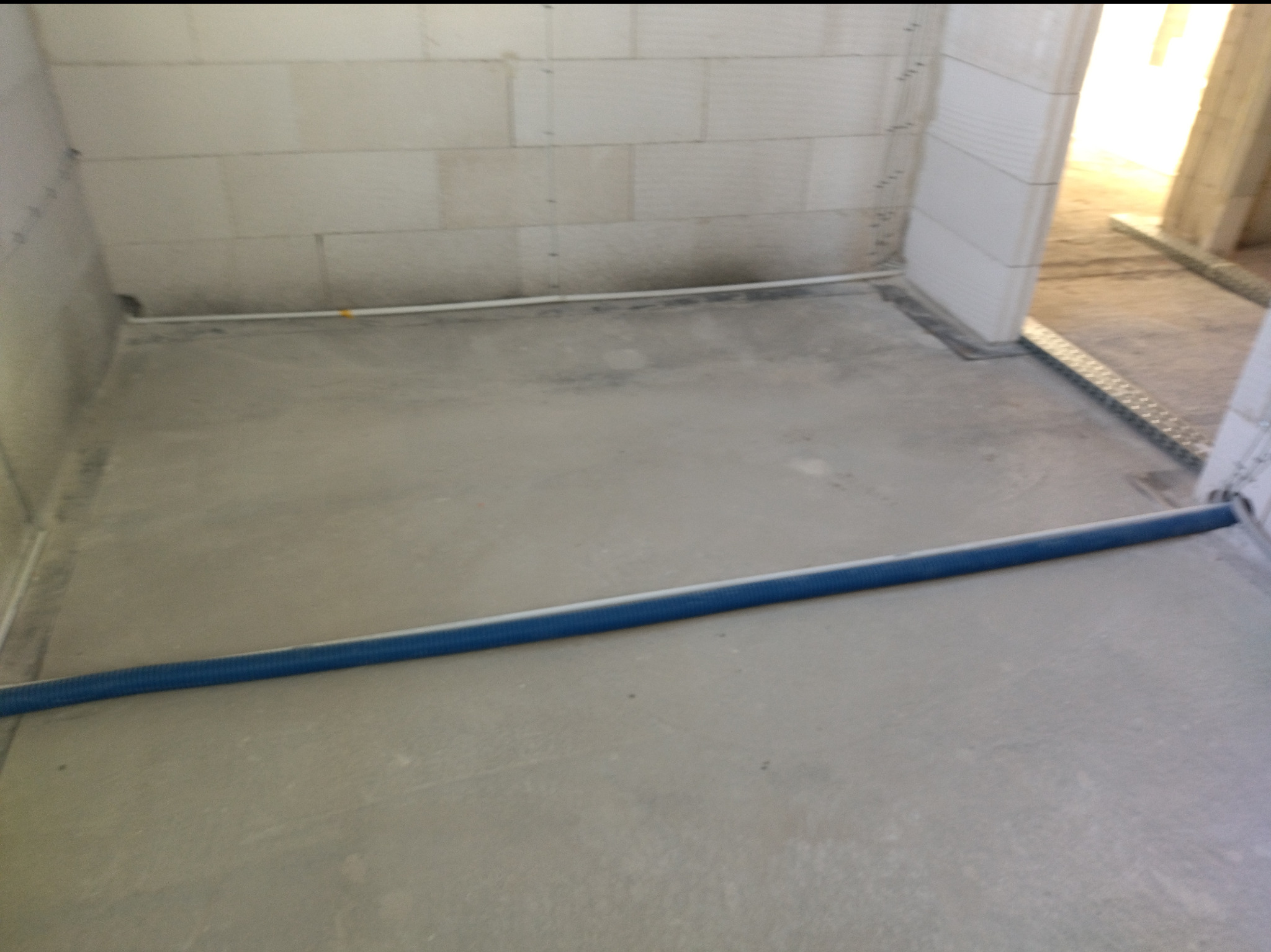 Wnętrze w trakcie prac: szara wylewka podłogowa, białe ściany z betonu komórkowego, niebieska rura instalacyjna wzdłuż progu. Widoczne elementy instalacji elektrycznej.