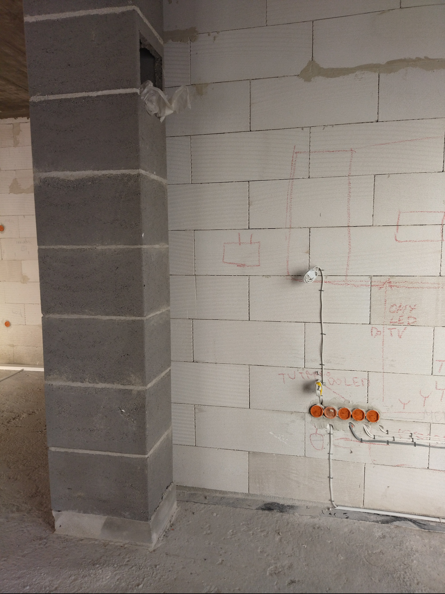 Ściana z betonu komórkowego z instalacją elektryczną w trakcie montażu: puszki pod gniazdka i przewody. Na ścianie widoczne szkice rozplanowania instalacji.