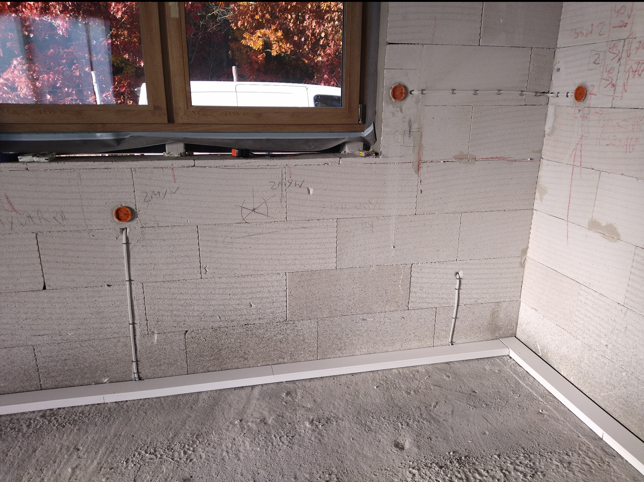 Surowy stan: ściana z betonu komórkowego z puszkami elektrycznymi i pionowymi bruzdami na przewody, listwa przypodłogowa, okno z widokiem na drzewa.