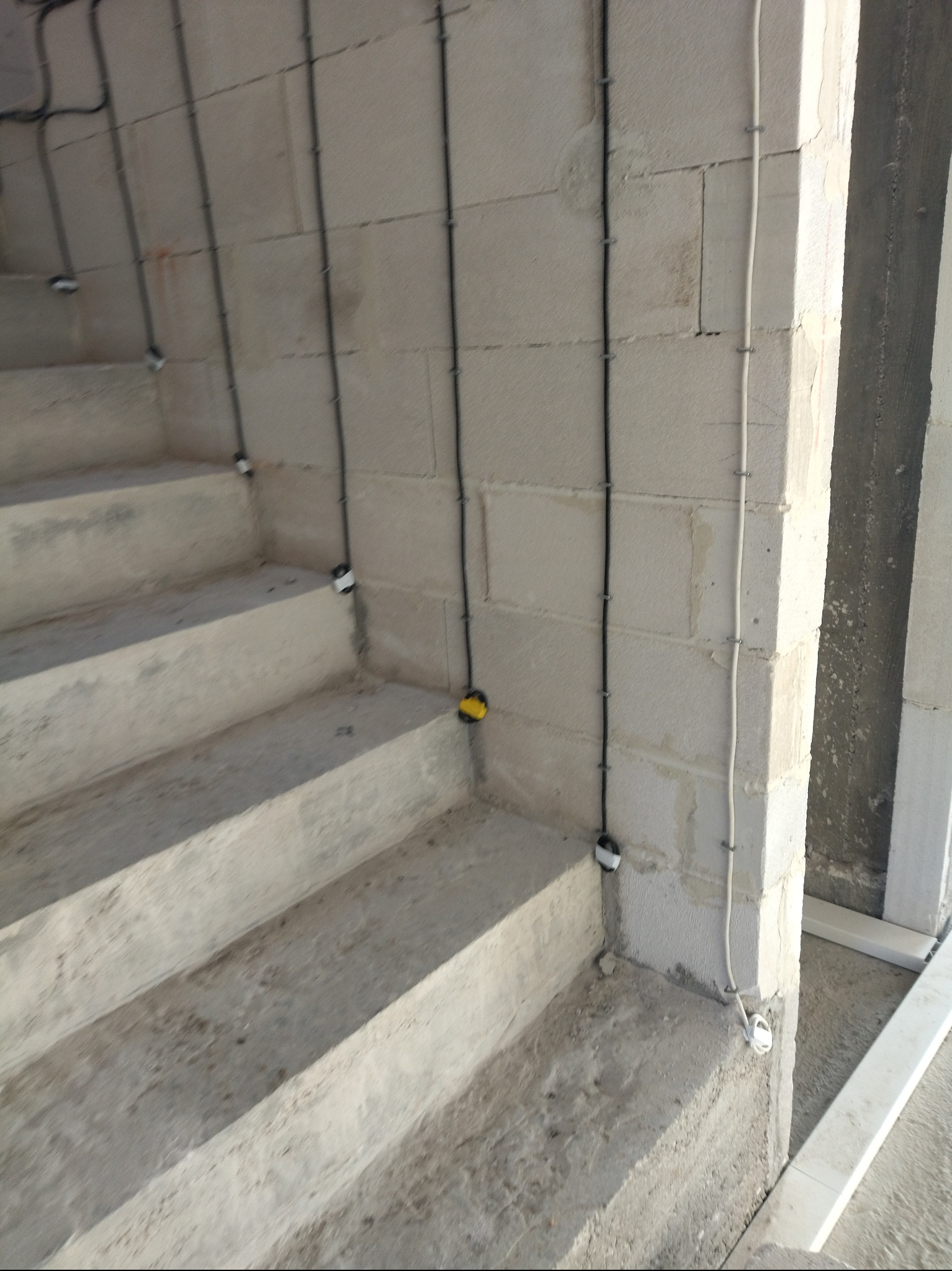 Kable elektryczne prowadzone wzdłuż surowych schodów z betonu komórkowego, zabezpieczone uchwytami. Widoczne puszki instalacyjne i przewody w kolorze białym i czarnym.