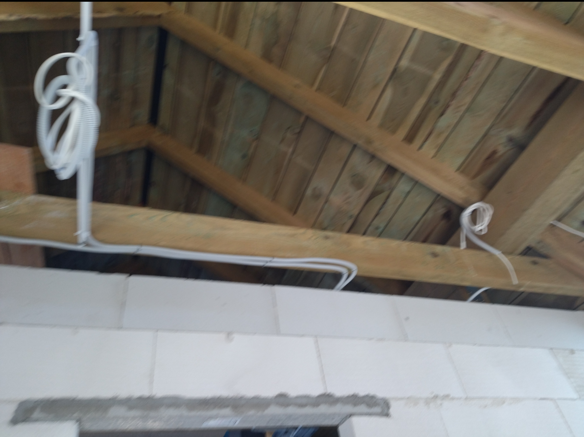 Białe przewody elektryczne prowadzone pod drewnianą konstrukcją dachu w surowym stanie, widoczne białe bloczki ścian. Instalacja w trakcie realizacji.