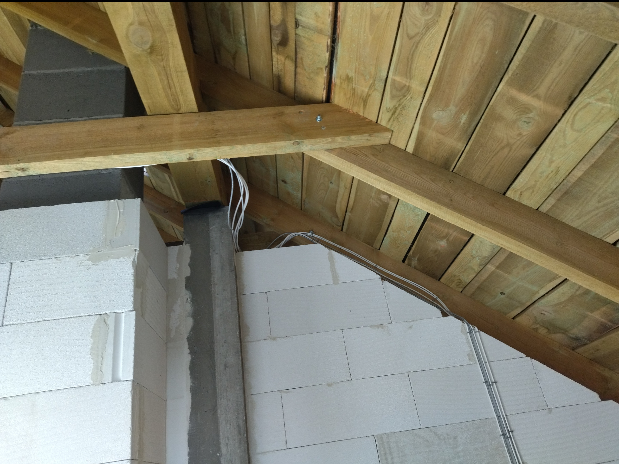 Surowy stan poddasza z instalacją kabli w Sząbruk. Widoczne drewniane elementy konstrukcyjne dachu i murowany komin. Kable prowadzone wzdłuż ściany z bloczków.