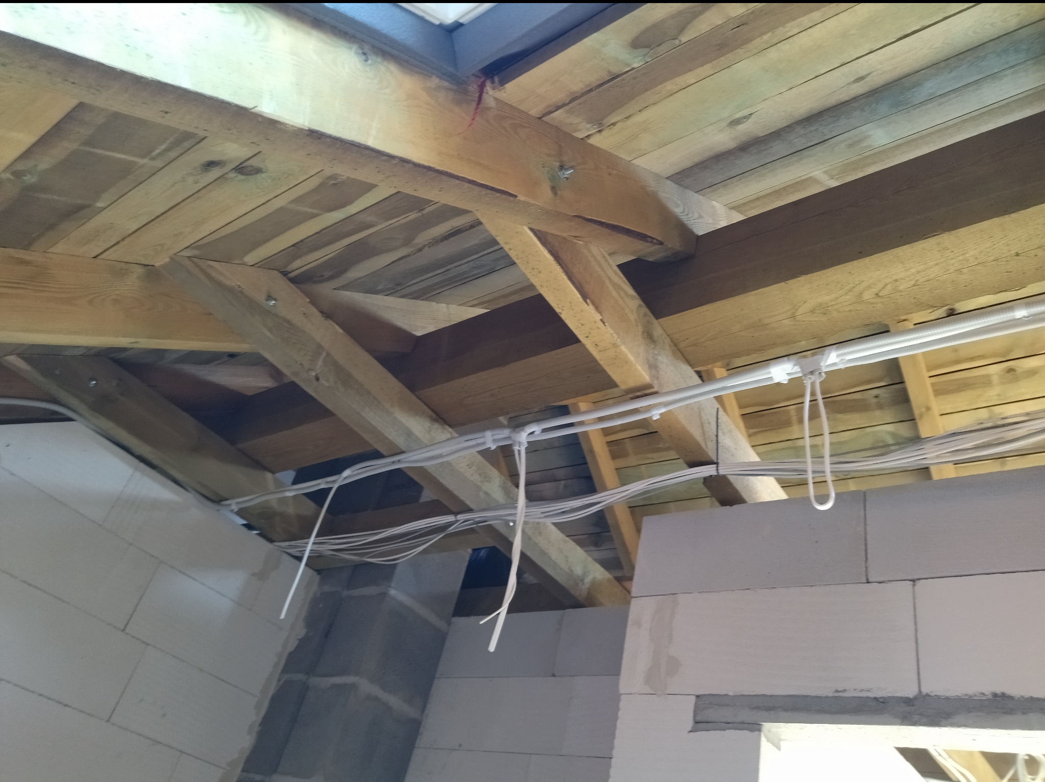 Białe kable elektryczne przymocowane do drewnianej konstrukcji dachu w surowym wnętrzu, widok z dołu na instalację na tle bloczków betonowych.