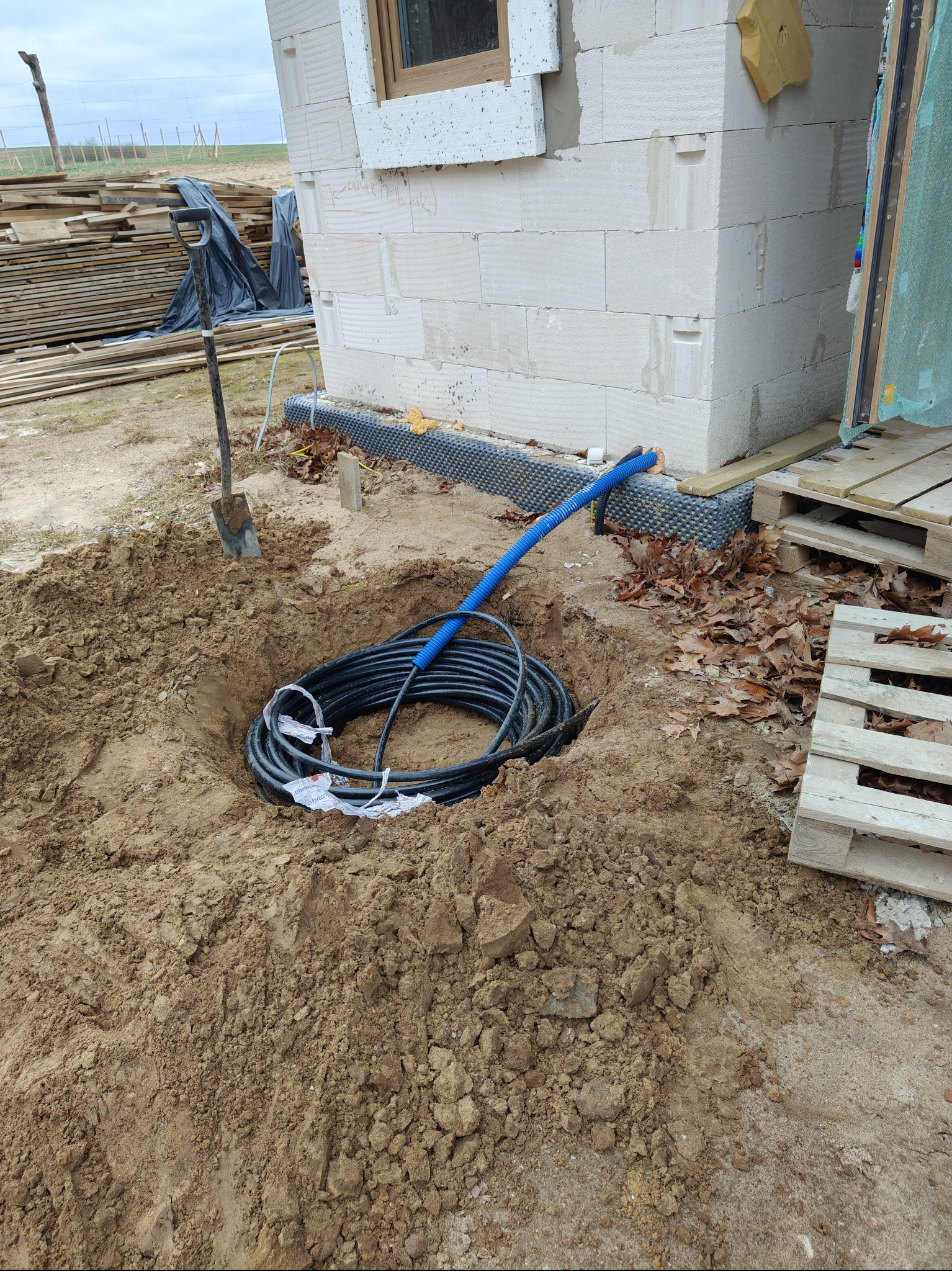 Kabel elektryczny w wykopie przy fundamencie budynku z betonu komórkowego. Widoczna niebieska rura osłonowa i szpadel. Etap prac ziemnych przed zasypaniem.