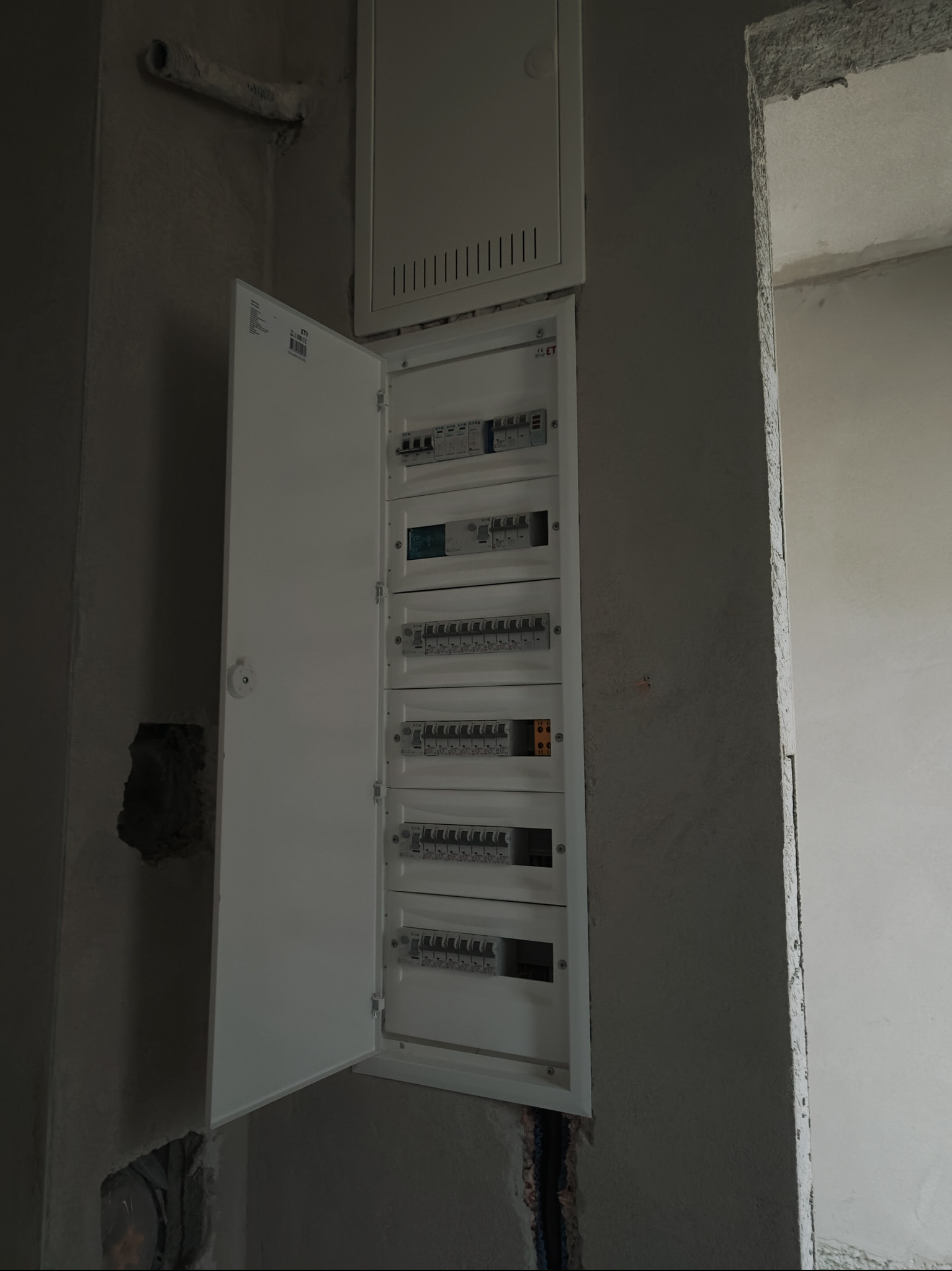 Otwarta skrzynka elektryczna z bezpiecznikami w surowym wnętrzu, widoczne przewody. Instalacja w trakcie prac wykończeniowych, ściany w stanie deweloperskim.