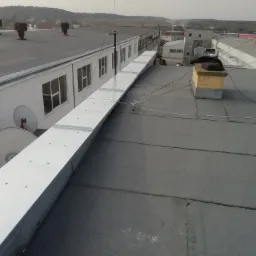 Widok z góry na płaski dach pokryty papą z nową, długą, metalową obróbką blacharską wzdłuż ściany budynku z antenami satelitarnymi.