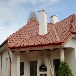 Nowy, błyszczący dach z czerwonej dachówki ceramicznej na jasnym domu jednorodzinnym, widok z dołu, z widocznymi kominami i rynnami w kolorze dachu.