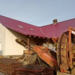 Dom z nowym dachem z blachy trapezowej w kolorze burgundowym, na pierwszym planie sterta starych dachówek i maszyny rolnicze.