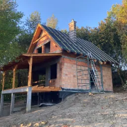 Niedokończony dom z czerwonej cegły z drewnianą konstrukcją dachu i kominem, widoczne rusztowanie i teren budowy w otoczeniu drzew.