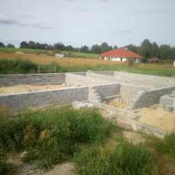 Fundamenty domu z bloczków betonowych na piaszczystym podłożu, widok z zewnątrz, w tle budynek mieszkalny z czerwoną dachówką i zarośnięte pole.
