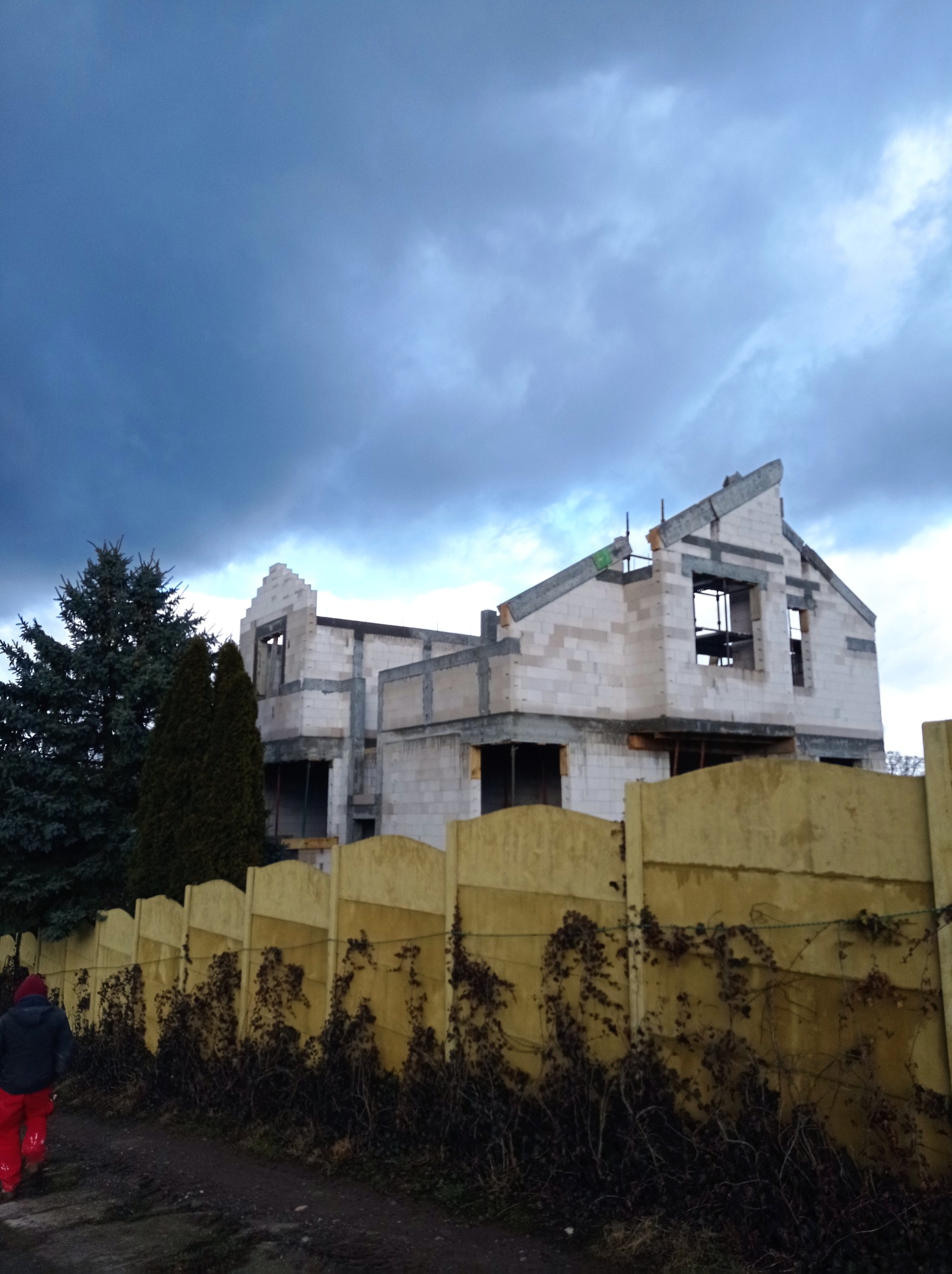 Surowy stan dwupiętrowego domu z pustaków, z widocznymi otworami okiennymi i dachem w trakcie budowy, sfotografowany zza żółtego ogrodzenia z pnączami na tle pochmurnego nieba i zielonych drzew.