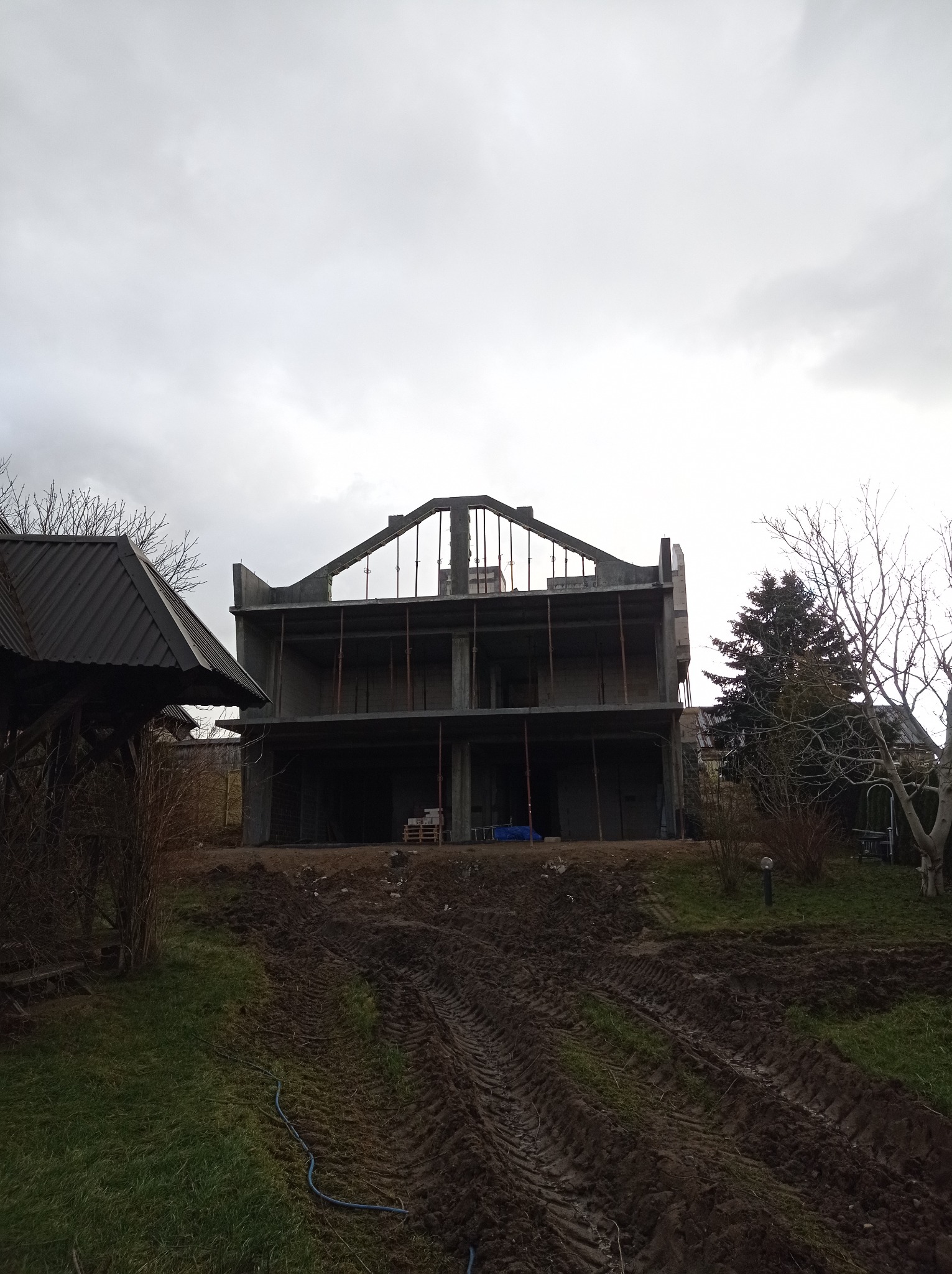 Dwukondygnacyjny dom w stanie surowym otwartym z widocznymi elementami konstrukcyjnymi, na pierwszym planie błotniste ślady opon i trawa, pochmurne niebo w tle.