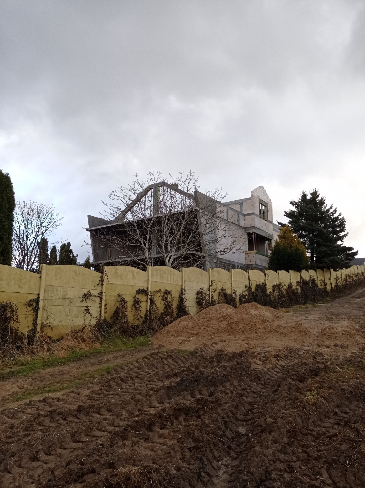 Niedokończony, dwupiętrowy dom w stanie surowym za żółtym, betonowym ogrodzeniem z pnączami, widok z dołu na tle pochmurnego nieba.