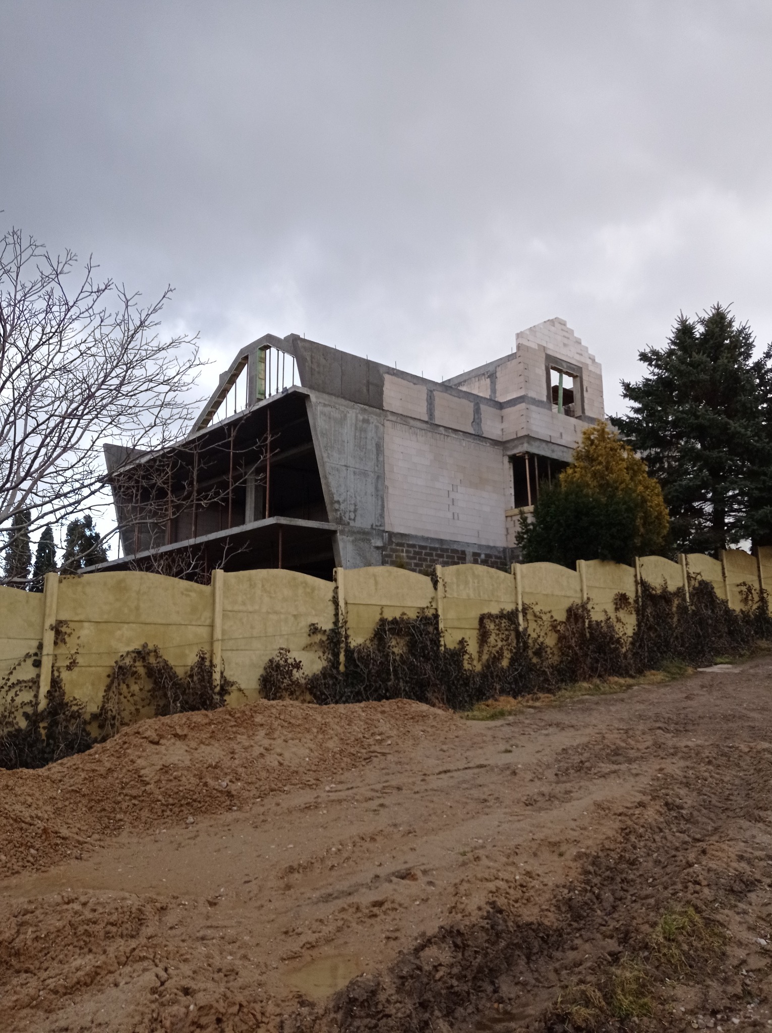 Surowa bryła domu w trakcie rozbudowy, z widocznymi elementami konstrukcyjnymi i pustakami, ogrodzona żółtym płotem, na tle pochmurnego nieba i drzew.