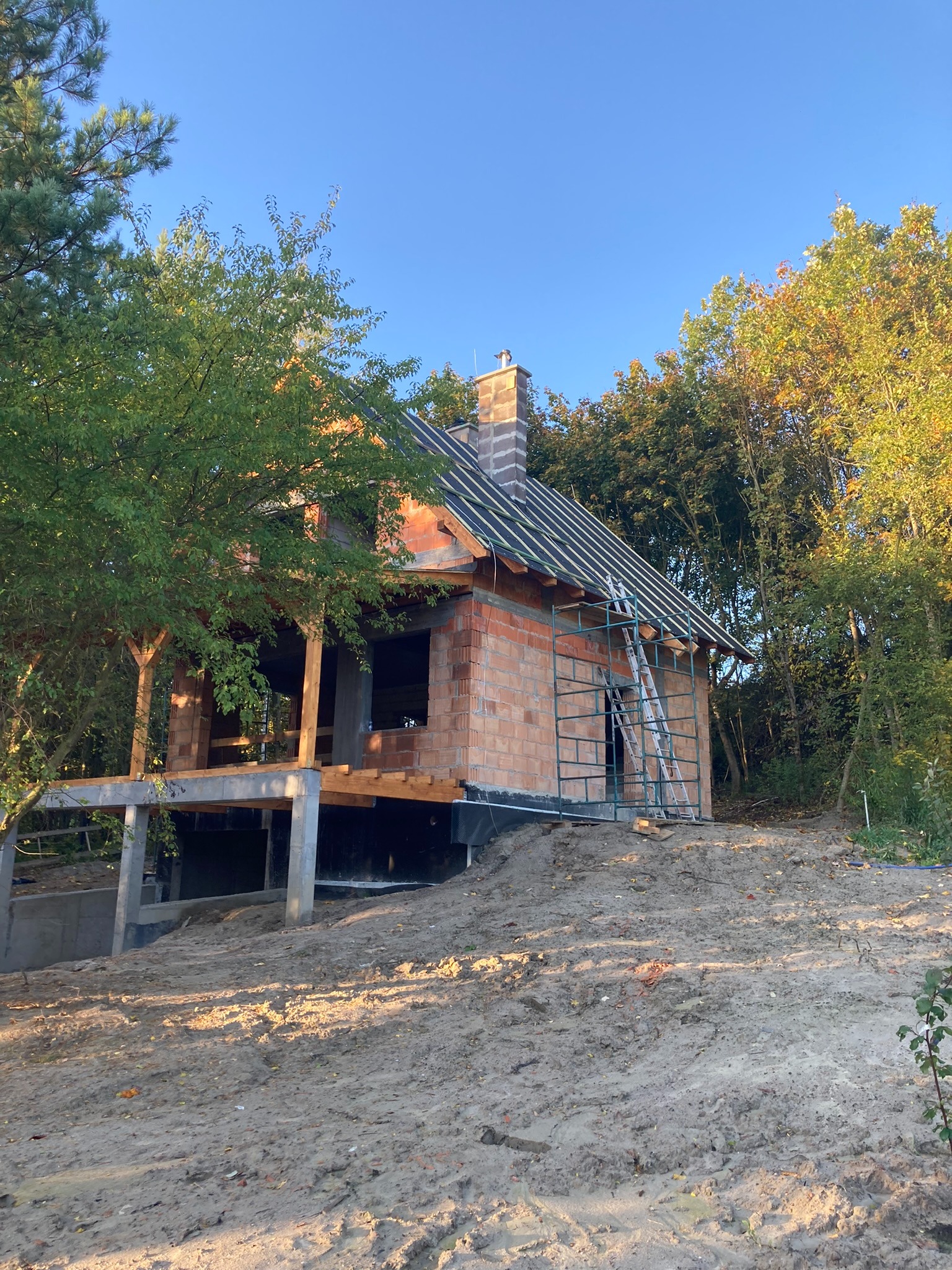 Dom w trakcie budowy z czerwonej cegły, widoczne rusztowanie i drabina przy ścianie, otoczony drzewami na wzniesieniu.