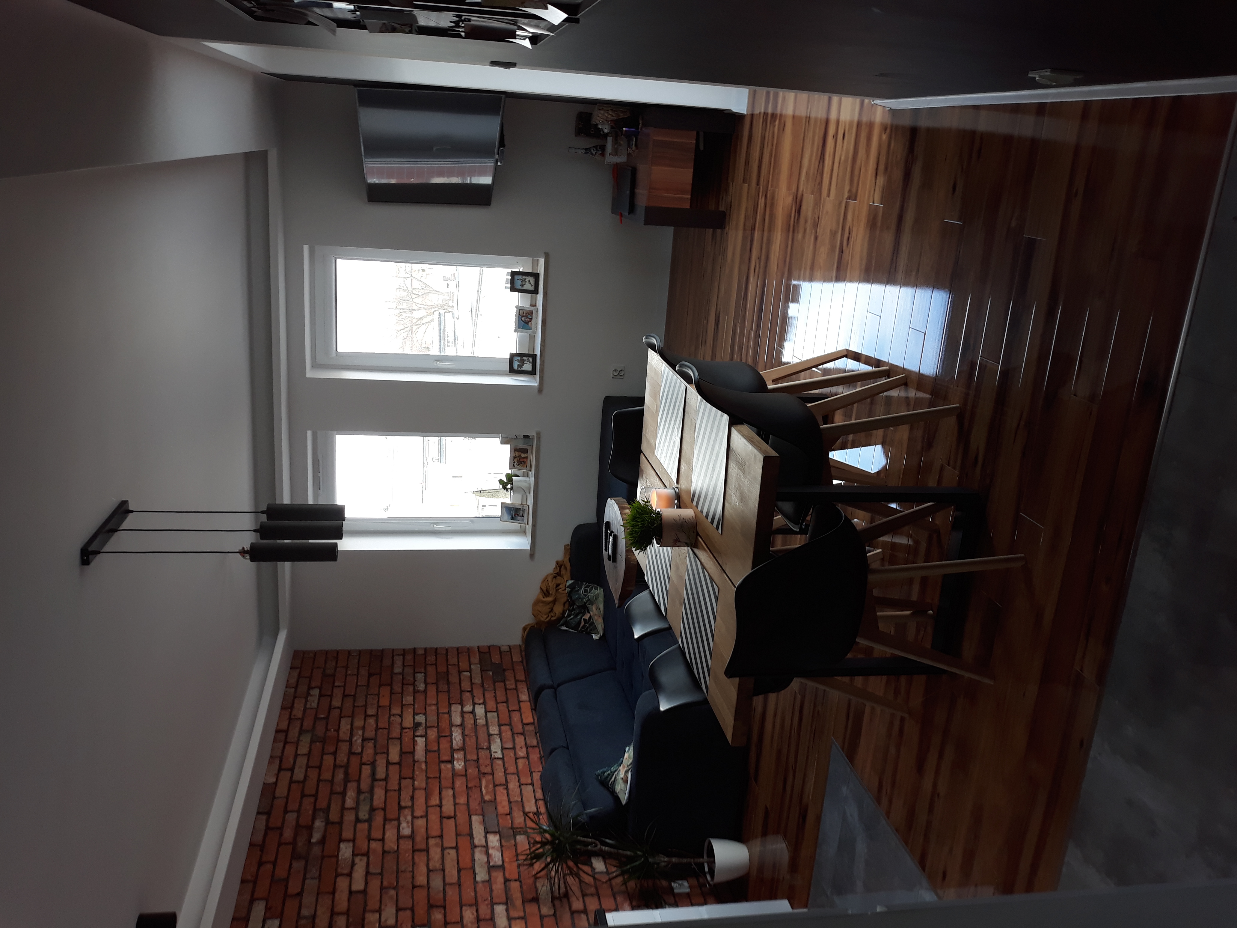 Jasne wnętrze z drewnianą podłogą, ceglaną ścianą, dwoma oknami i drewnianym stołem z czarnymi krzesłami, oświetlone wiszącą lampą.