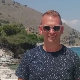 Uśmiechnięty mężczyzna w okularach przeciwsłonecznych na tle górzystego krajobrazu z widokiem na morze.