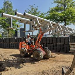 Marcin Olszewski - Operator w kasku przewozi pomarańczową mini-ładowarką konstrukcję ze srebrnych rur z przyspawanymi podstawami, tło stanowią ułożone palety z kostką brukową i budynek.