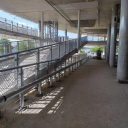 Marcin Olszewski - Metalowe barierki i poręcze na rampie dla pieszych w trakcie budowy, z widocznymi wystającymi śrubami mocującymi i betonowymi kolumnami podtrzymującymi konstrukcję.