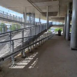 Metalowe barierki i poręcze na rampie dla pieszych w trakcie budowy, z widocznymi wystającymi śrubami mocującymi i betonowymi kolumnami podtrzymującymi konstrukcję.