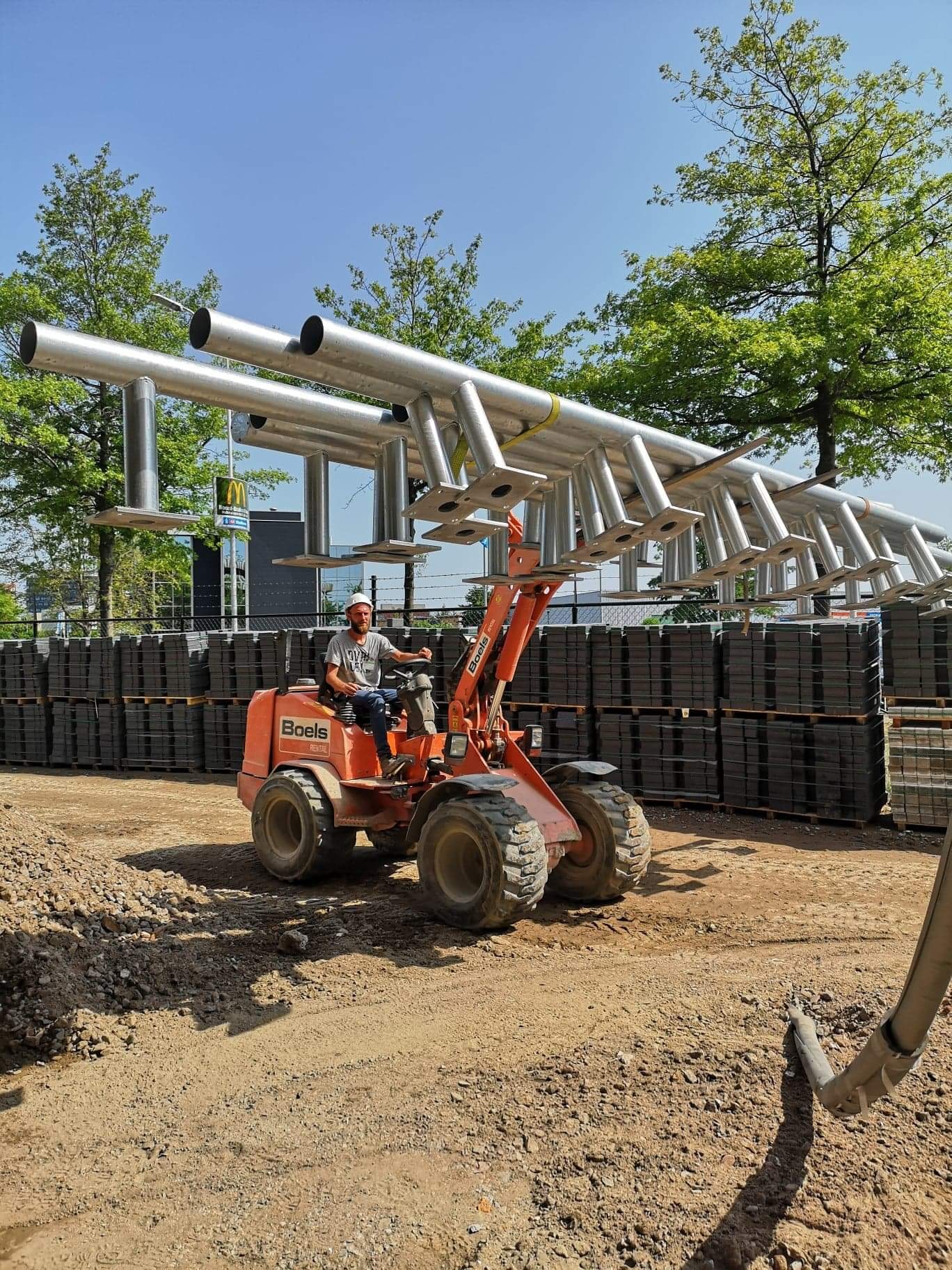 Operator w kasku przewozi pomarańczową mini-ładowarką konstrukcję ze srebrnych rur z przyspawanymi podstawami, tło stanowią ułożone palety z kostką brukową i budynek.