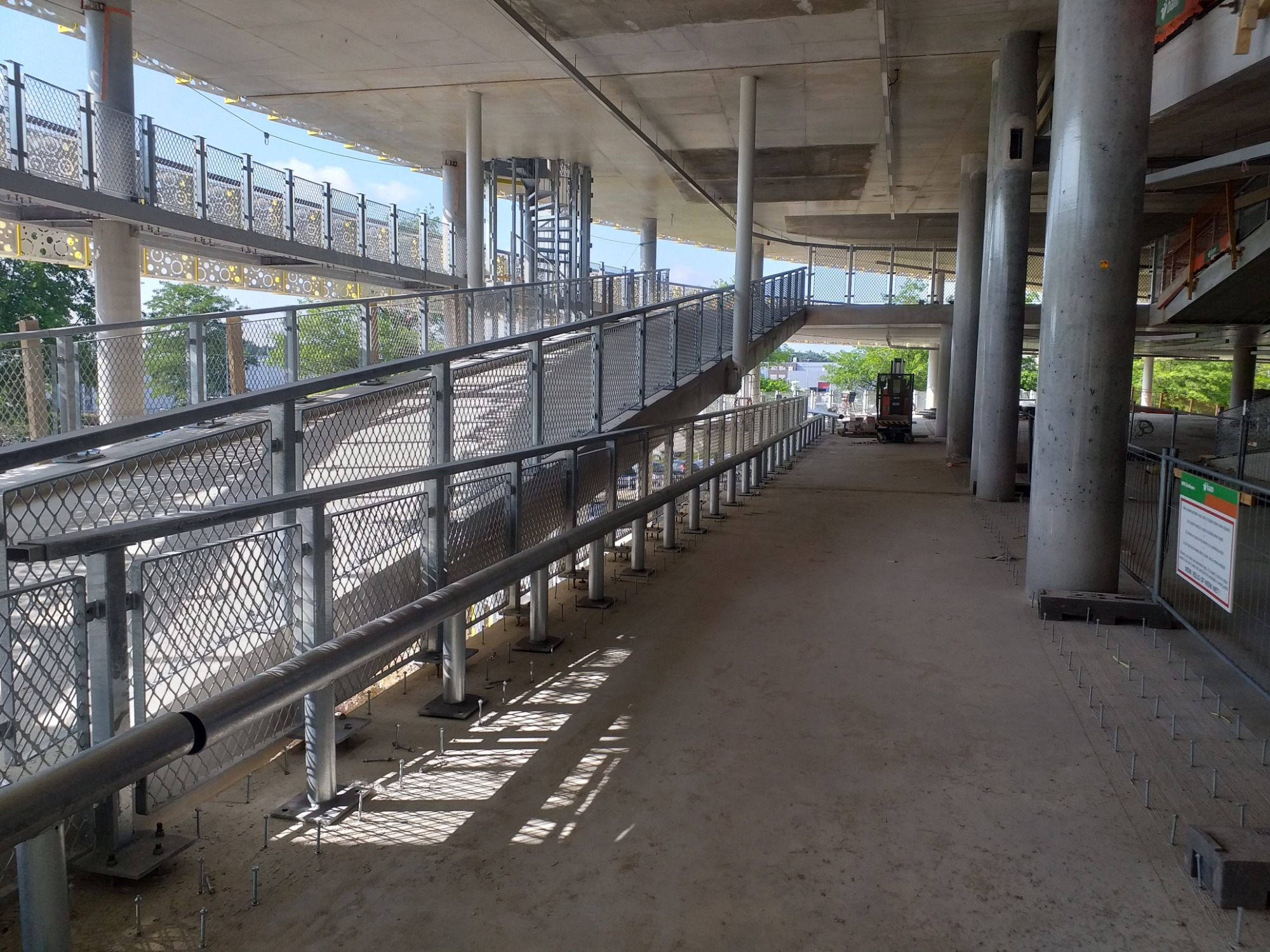 Metalowe barierki i poręcze na rampie dla pieszych w trakcie budowy, z widocznymi wystającymi śrubami mocującymi i betonowymi kolumnami podtrzymującymi konstrukcję.