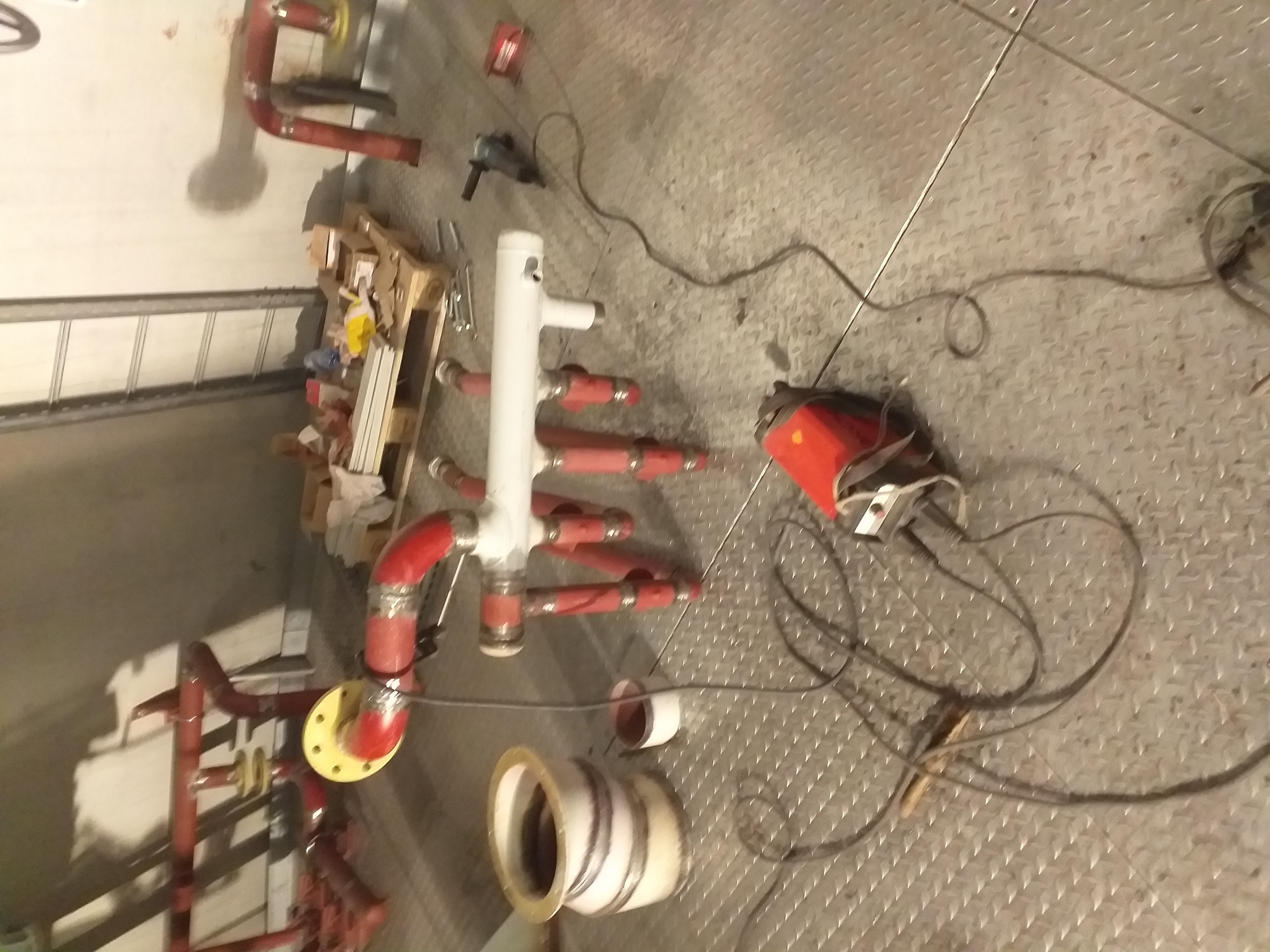 Warsztatowa podłoga z blachy ryflowanej, na której leżą elementy instalacji rurowej w kolorze czerwonym i białym, spawarka z przewodami, paleta i elektronarzędzie.