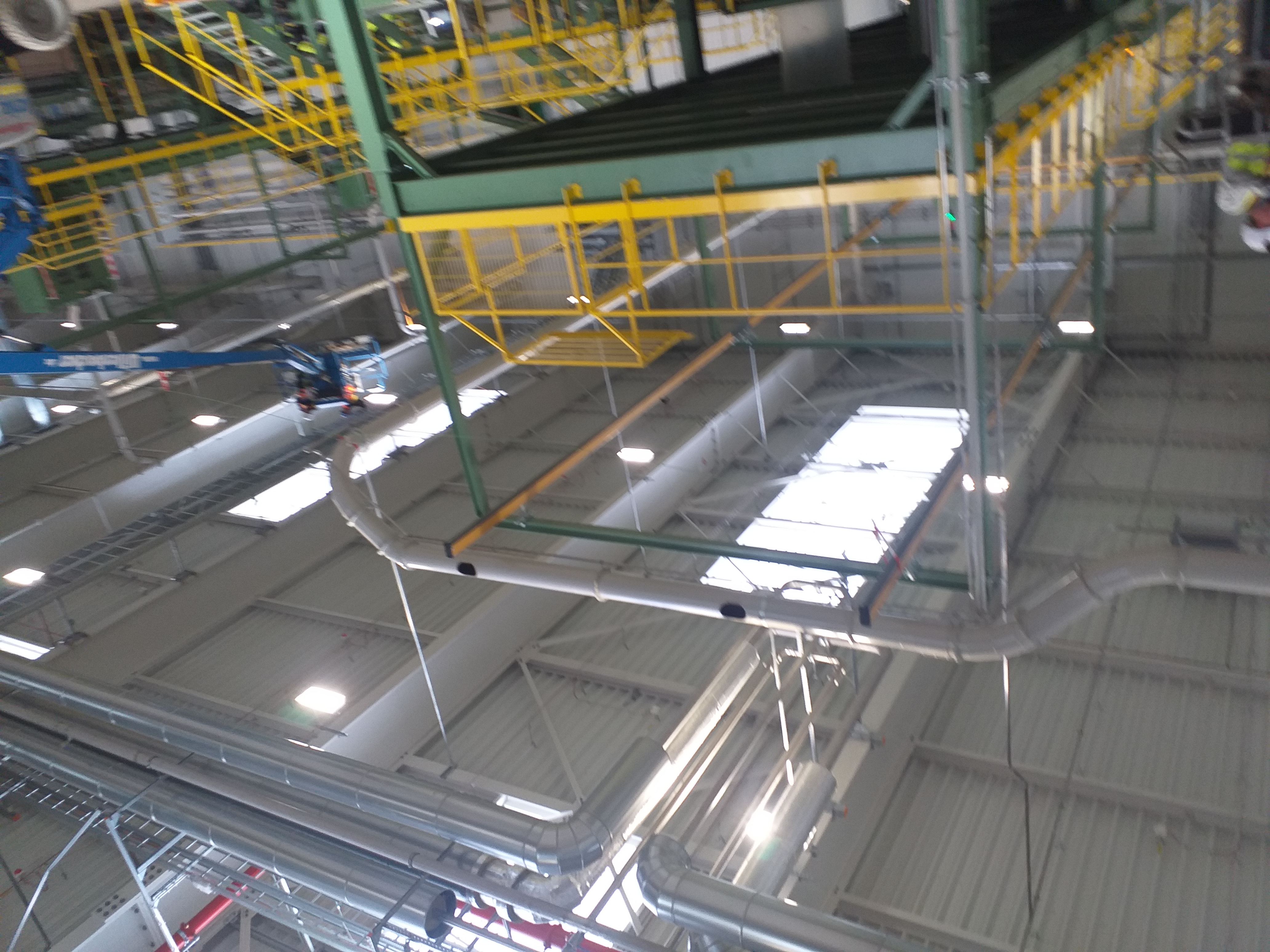 Wysoka hala przemysłowa z widoczną konstrukcją stalową, rurami wentylacyjnymi i platformą roboczą na wysokości, oświetlona światłem dziennym wpadającym przez okna dachowe.