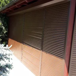 Zewnętrzny widok na brązowe rolety garażowe z metalowymi lamelami, zamontowane na ścianie budynku z drewnianymi elementami konstrukcyjnymi, widoczne fragmenty zieleni i niebieskiego nieba.